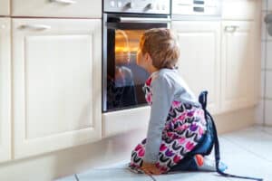 Cute funny blond kid boy baking muffins in domestic kitchen. Child having fun with helping, sitting near ofen and waiting for cupcakes.